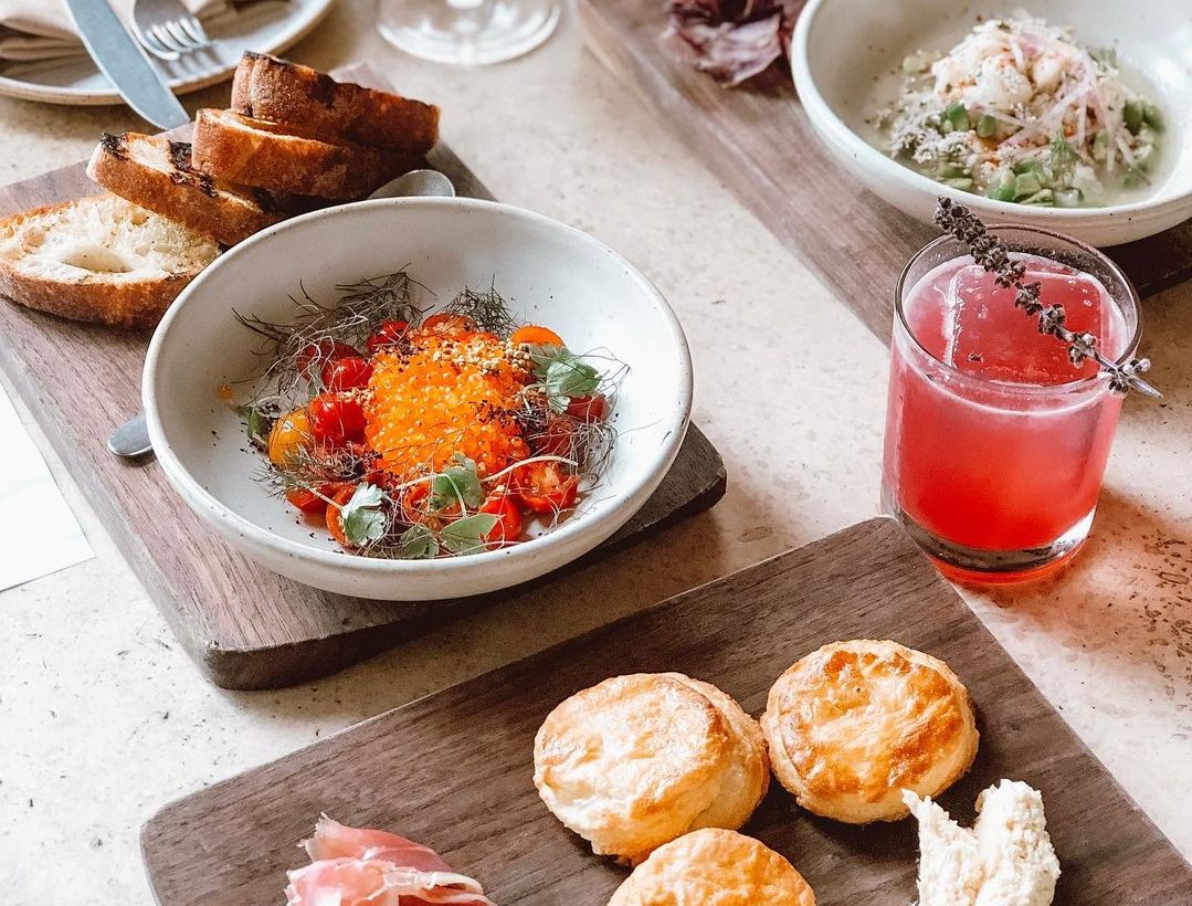 A table filled with a variety of dishes including a colorful tomato salad topped with orange roe, toasted bread slices, and a red cocktail garnished with lavender.
