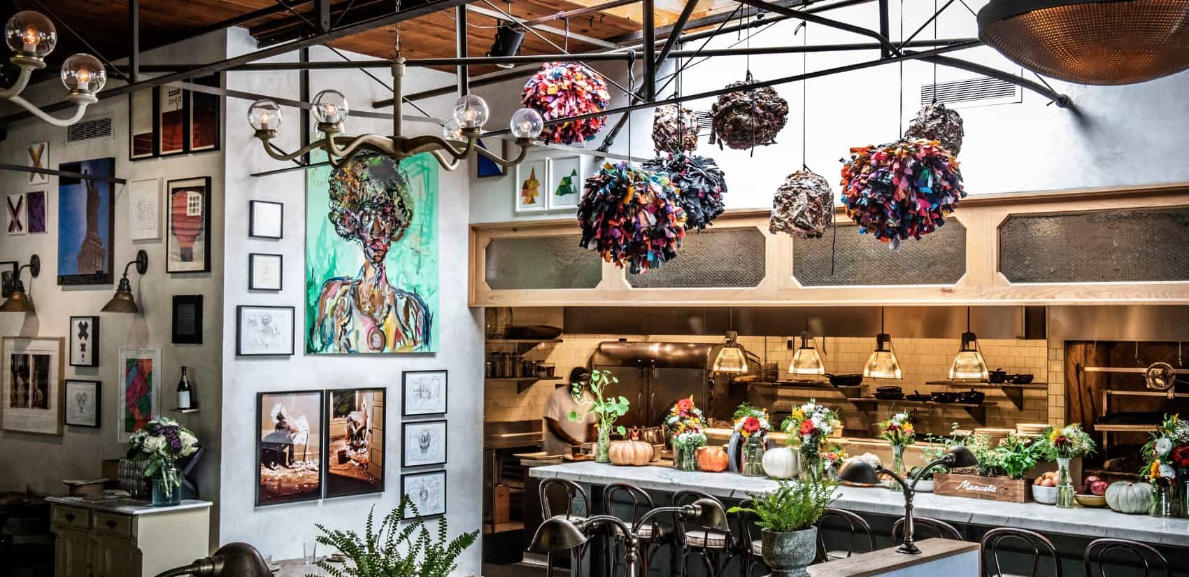 A vibrant restaurant interior featuring colorful art pieces on the wall, floral centerpieces, and a modern open kitchen with hanging decorative lights made from assorted fabric clusters.