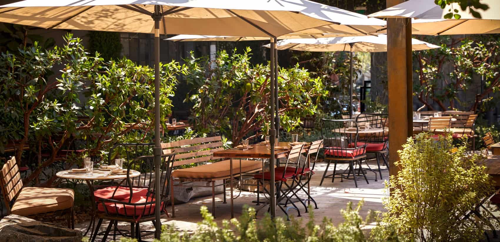 Outdoor patio seating area at a restaurant with shaded tables, red-cushioned chairs, and lush greenery surrounding the space.