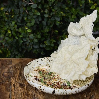A textured bowl of creamy dip garnished with herbs and spices, served with large, crispy white rice crackers.
