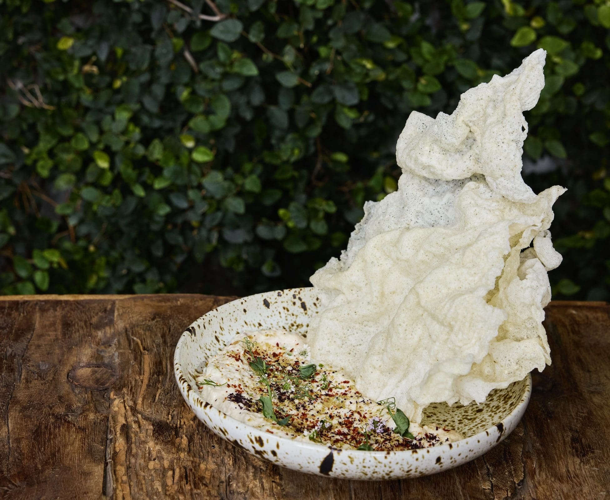 A textured bowl of creamy dip garnished with herbs and spices, served with large, crispy white rice crackers.