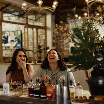 Two women laughing and enjoying drinks at a bar, with bottles and a vase of flowers in the foreground.