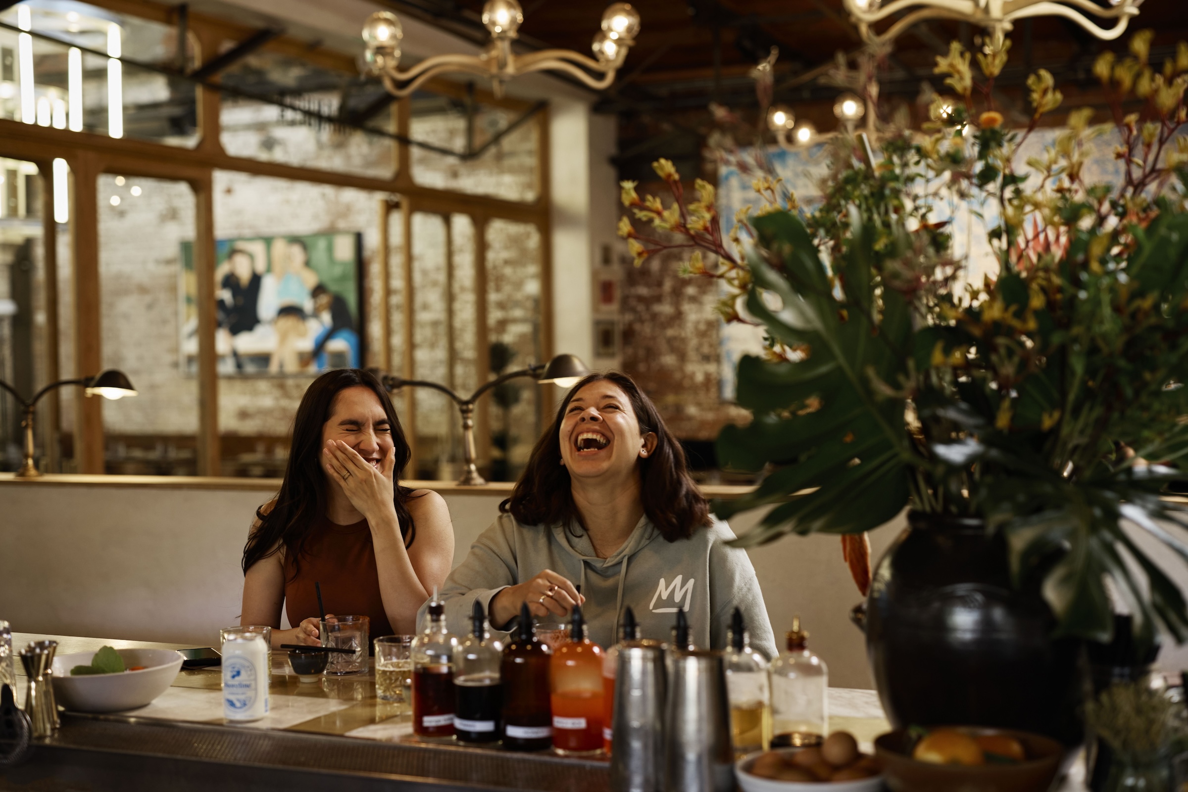 Two women laughing and enjoying drinks at a bar, with bottles and a vase of flowers in the foreground.