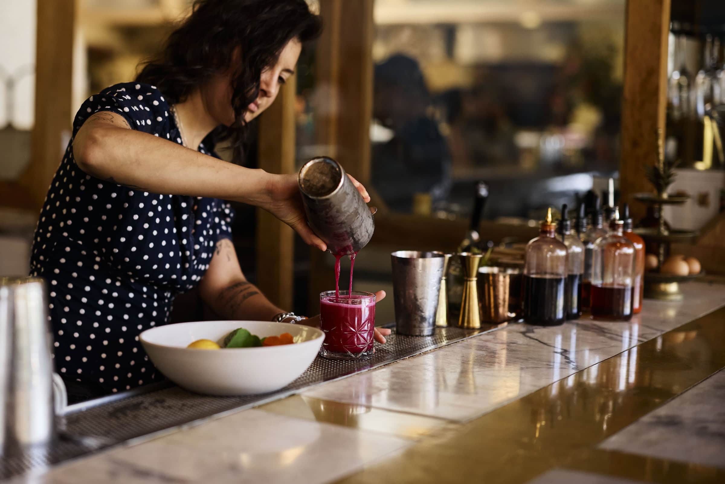 A bartender pouring a bright pink drink from a shaker into a glass at a bar, with bar tools and bottles in the background.