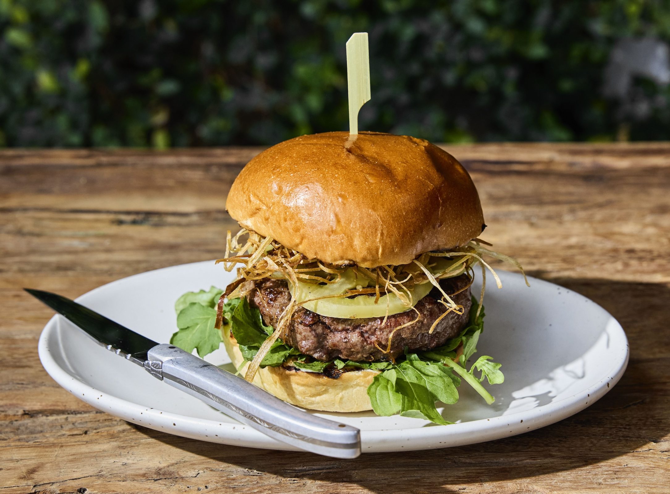 A beef burger topped with crispy fried onions, pickles, and greens, served on a white plate with a knife on the side.