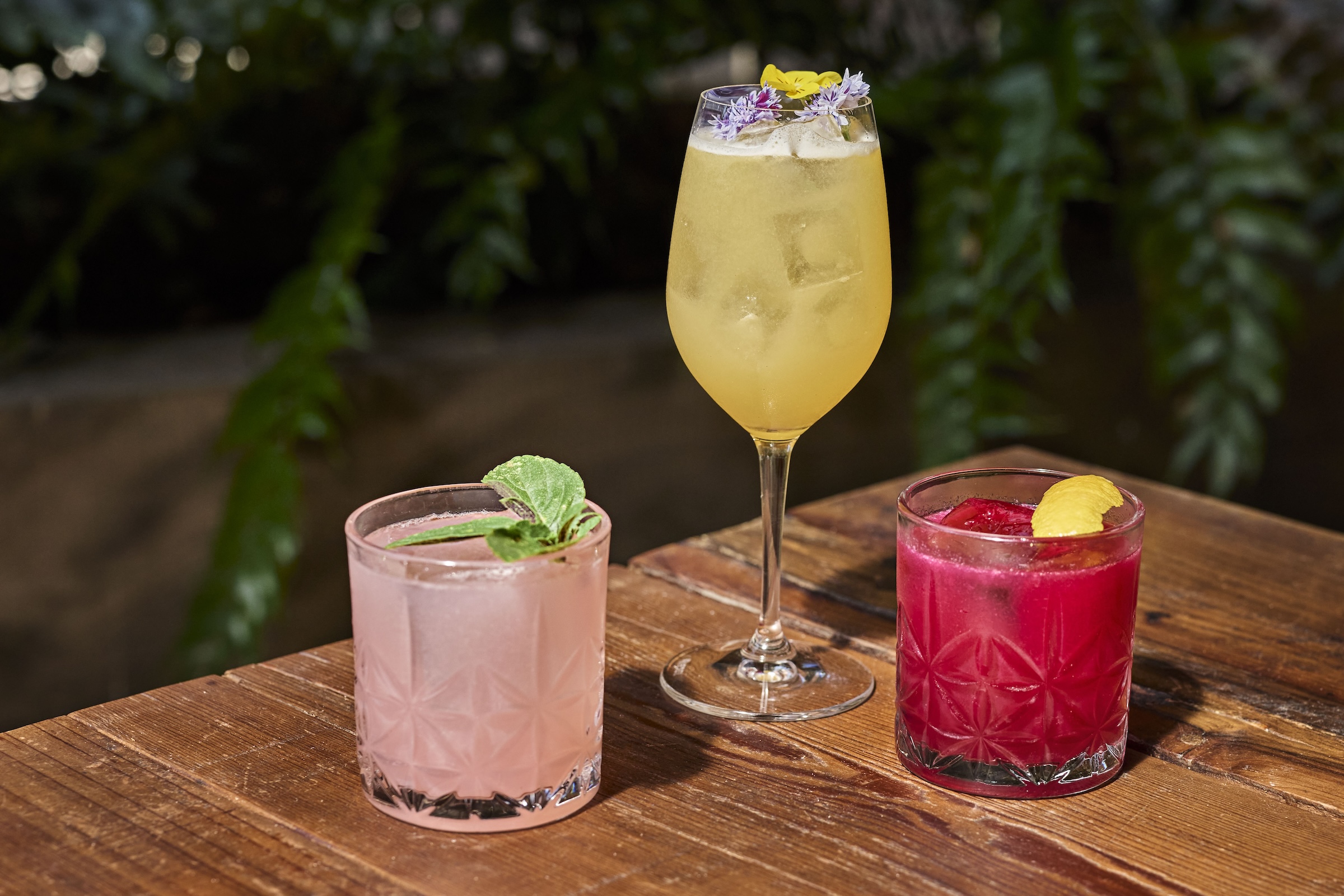 Three cocktails on a wooden table: a light pink drink with mint, a yellow drink in a wine glass garnished with flowers, and a bright red drink with a lemon peel.