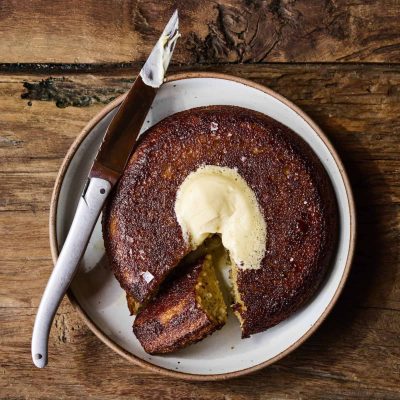 A round, golden-brown cornbread with a dollop of melting butter in the center, served on a plate with a knife beside it.