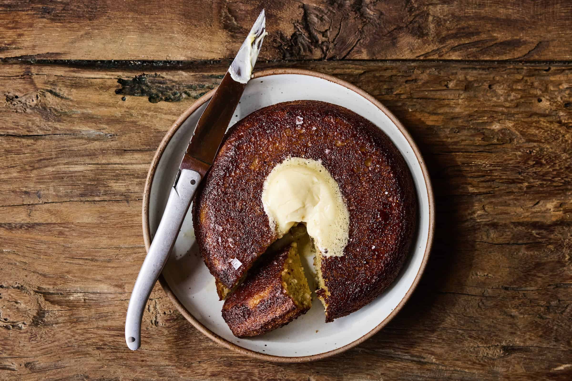 A round, golden-brown cornbread with a dollop of melting butter in the center, served on a plate with a knife beside it.