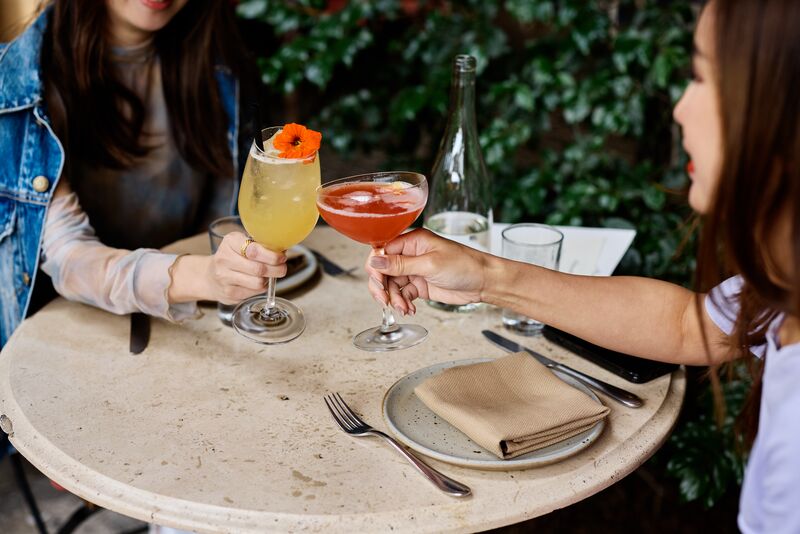 Two women clinking glasses with colorful cocktails at a round outdoor table set with plates, utensils, and a napkin.