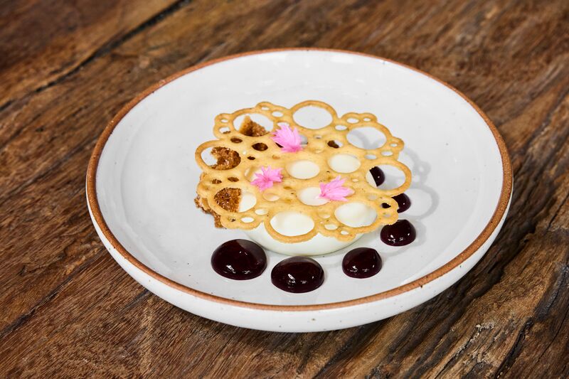 A plated goat's cheese cake dessert on a wood table. The dessert has a white bawl covered in a lace-like pastry surrounded by black dots.