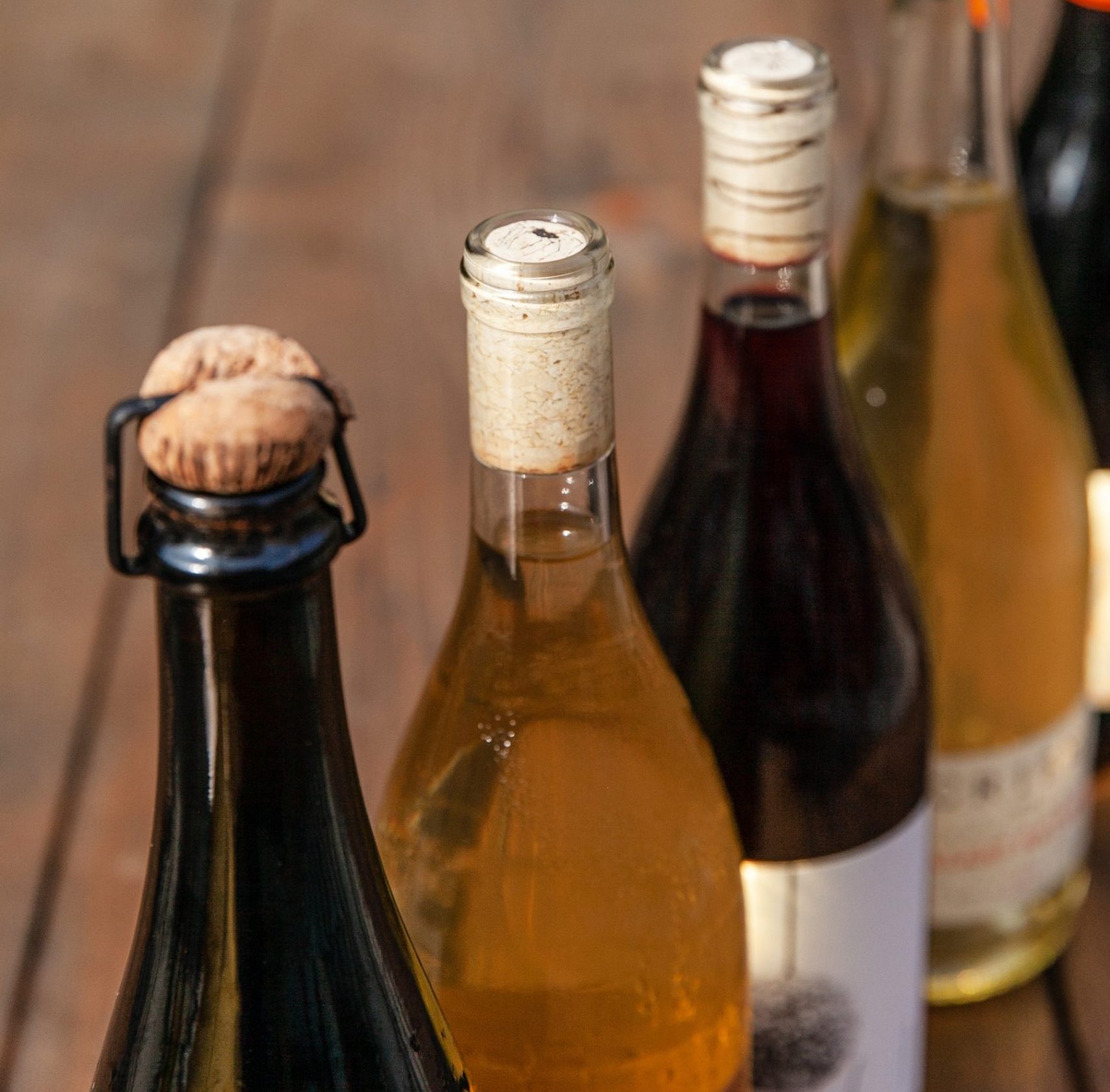 A close up of the tops of wine bottles lined up on a wooden table outside.