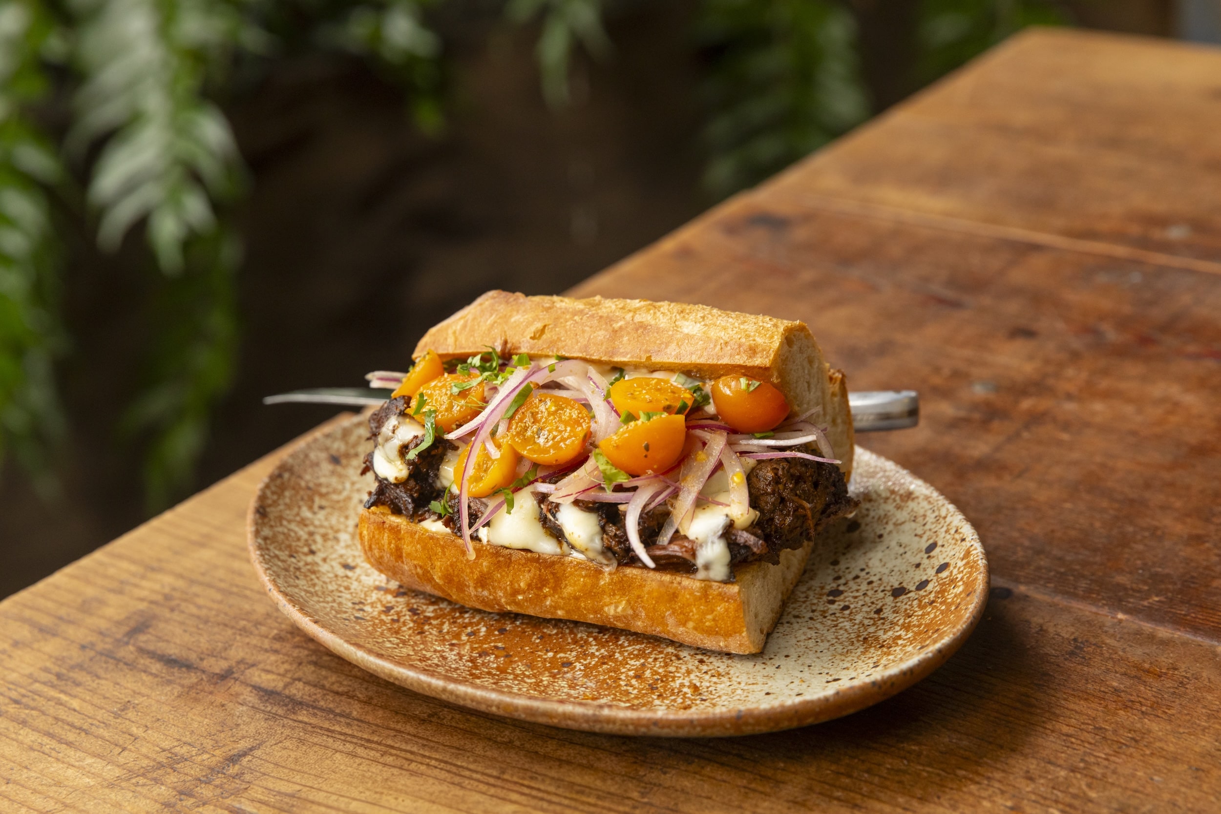 A baguette sandwhich full of shredded meat, melted cheese, sungold tomatoes and red onions on a brown textured plate. The dish is the Barbacoa sandwich.