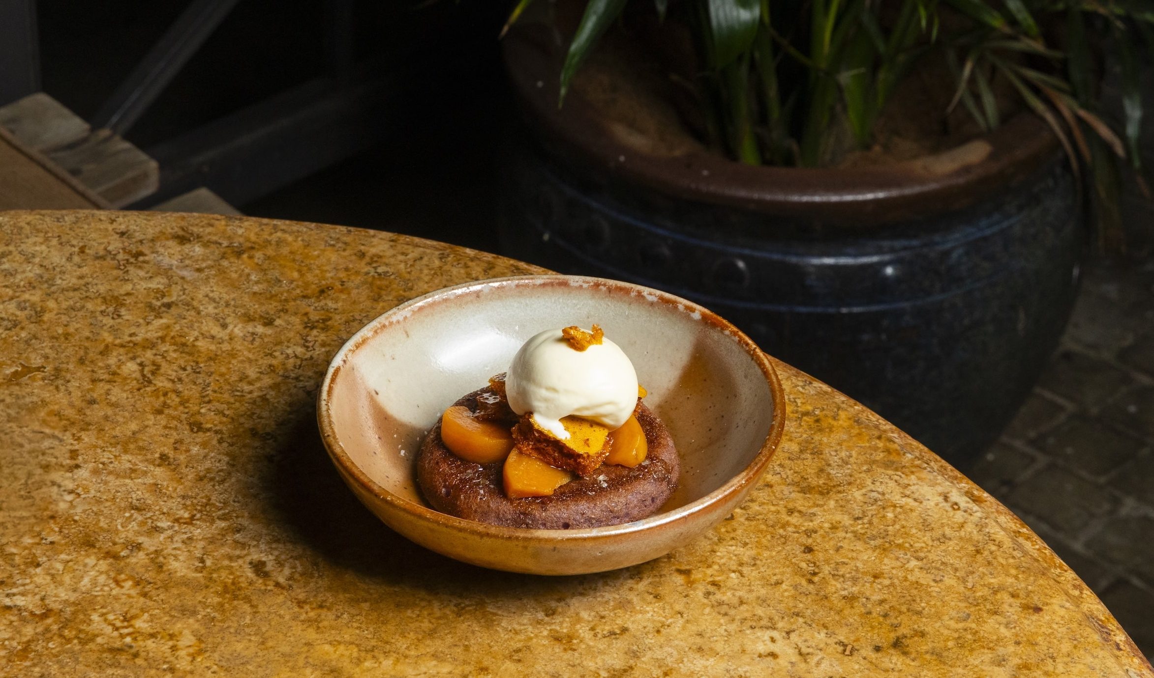 A dessert cake topped with peaches and white ice cream, served on a white and brown textured bowl.