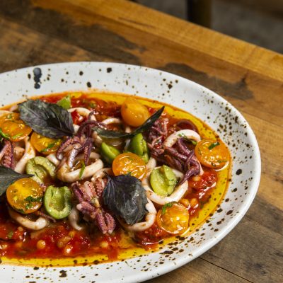 White plate with black spots holding a mixture of squid, tomatoes, olives, tomatoes and olive oil. The dish is Local squid with fregola.