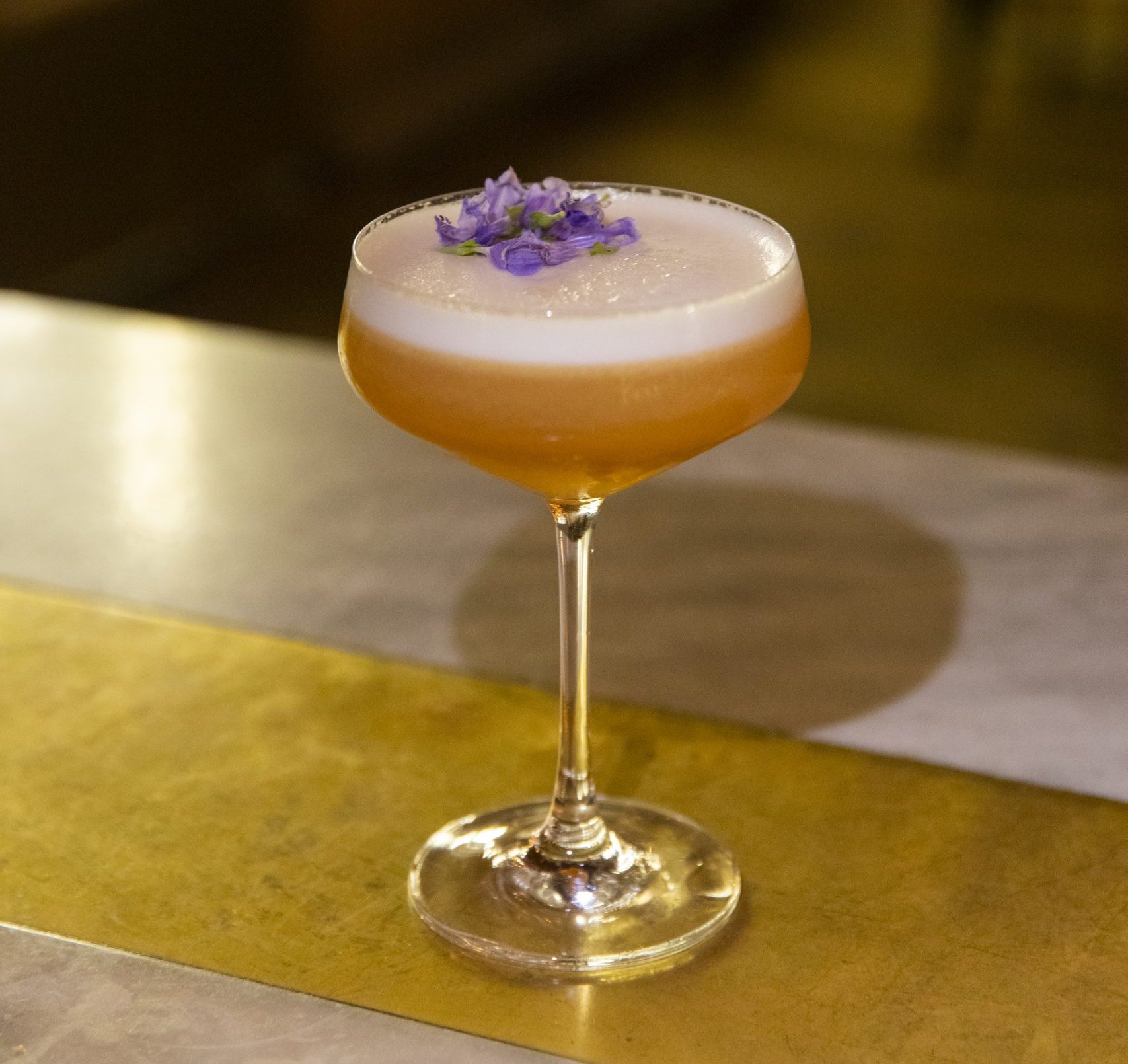 Brownish shaken cocktail in a coupe glass with purple flower garnish sitting on a gold and marble bar top.