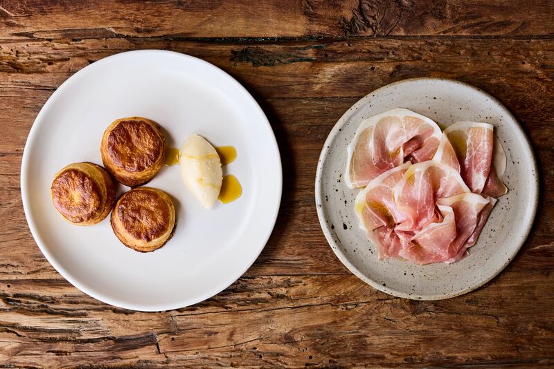 Three biscuits on a white plate with butter. A second textured plate with thin slices of ham sit next to it.