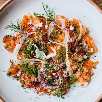 A white plate with cut up raw trout covered with sliced fennel, onion and capers. The dish is Ocean trout tartare.