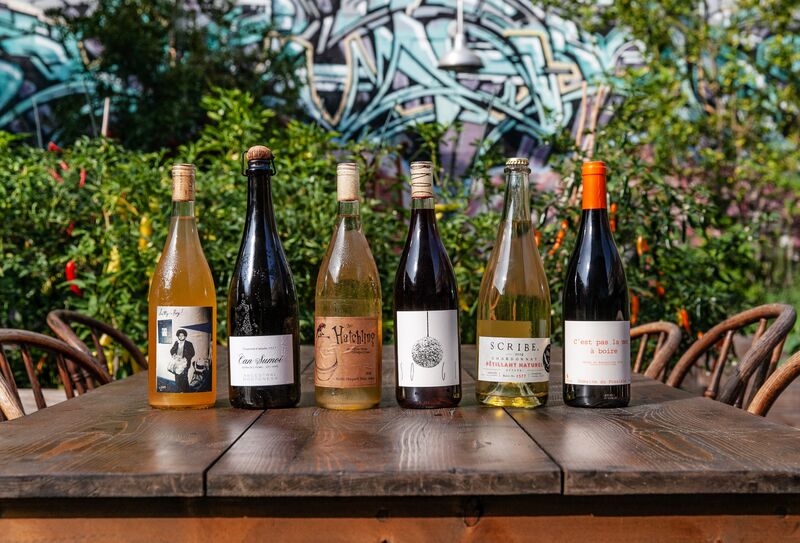 A row of six wine bottles sitting on a wooden table in with greenery in the background.