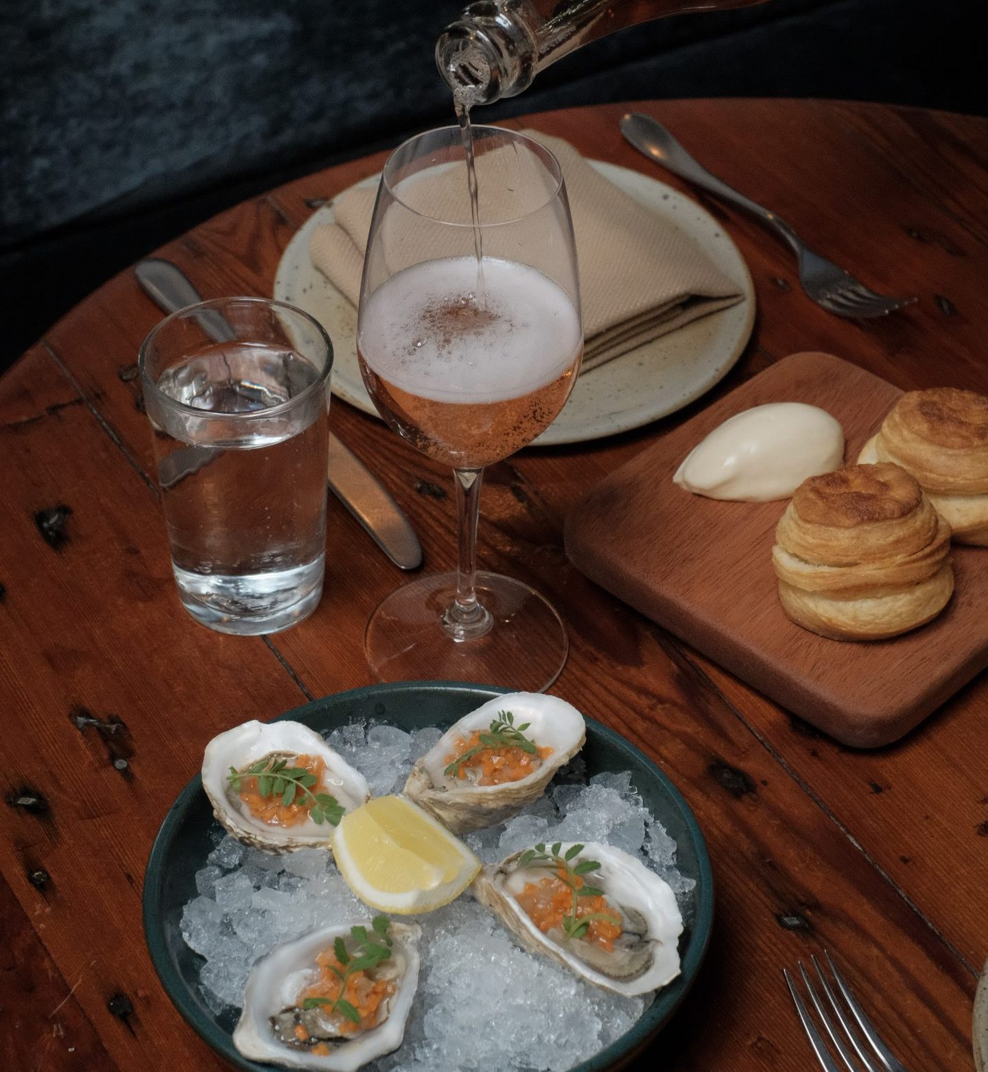 Pink sparkling wine being poured into a wine glass for Valentine's Day. Two plates of food are with it, one with oysters and the other with biscuits and butter.