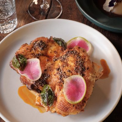 Baked half chicken on a white plate, garnished with green sauce, orange sauce, and slices of pink radishes.