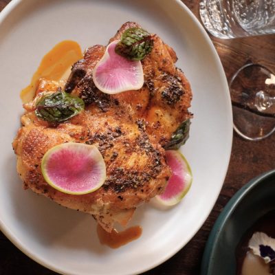 Baked half chicken on a white plate, garnished with green sauce, orange sauce, and slices of pink radishes.