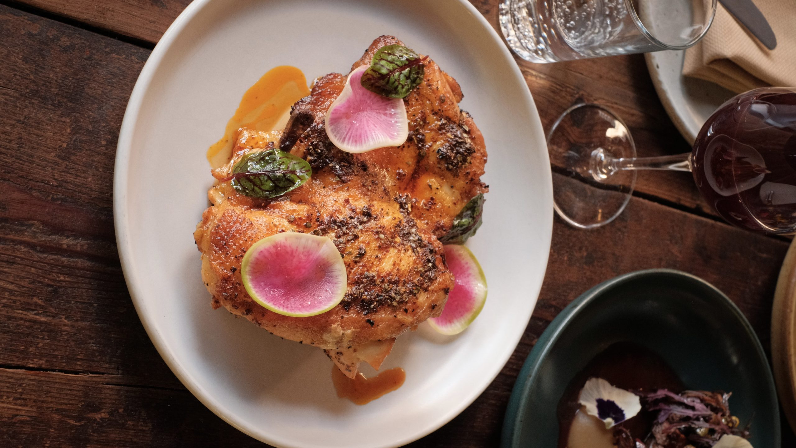 Baked half chicken on a white plate, garnished with green sauce, orange sauce, and slices of pink radishes.