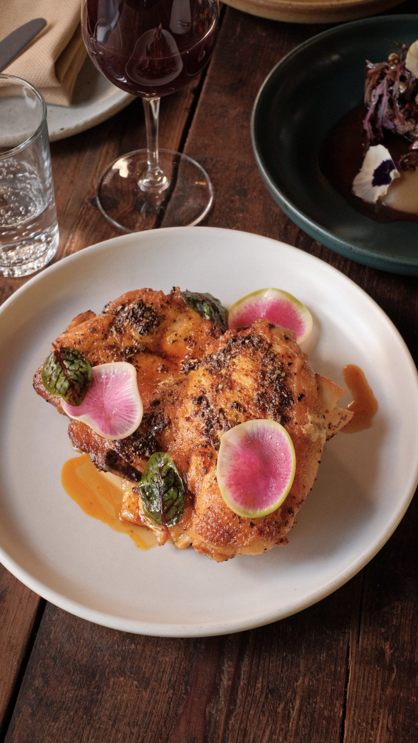 Baked half chicken on a white plate, garnished with green sauce, orange sauce, and slices of pink radishes.
