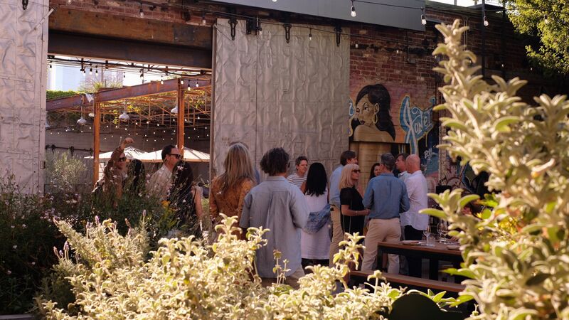 A group of people socializing in the garden area of Manuela in front of a large, sliding metal door. People are framed by light green bushes.