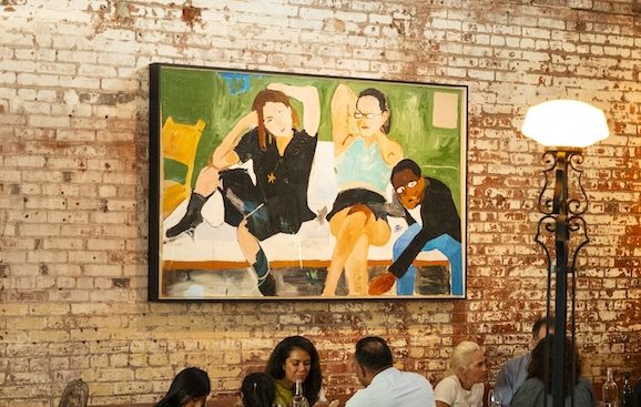 A colorful painting with blurred brush strokes of people talking together. The painting is hung on a brick wall with people sitting below it.