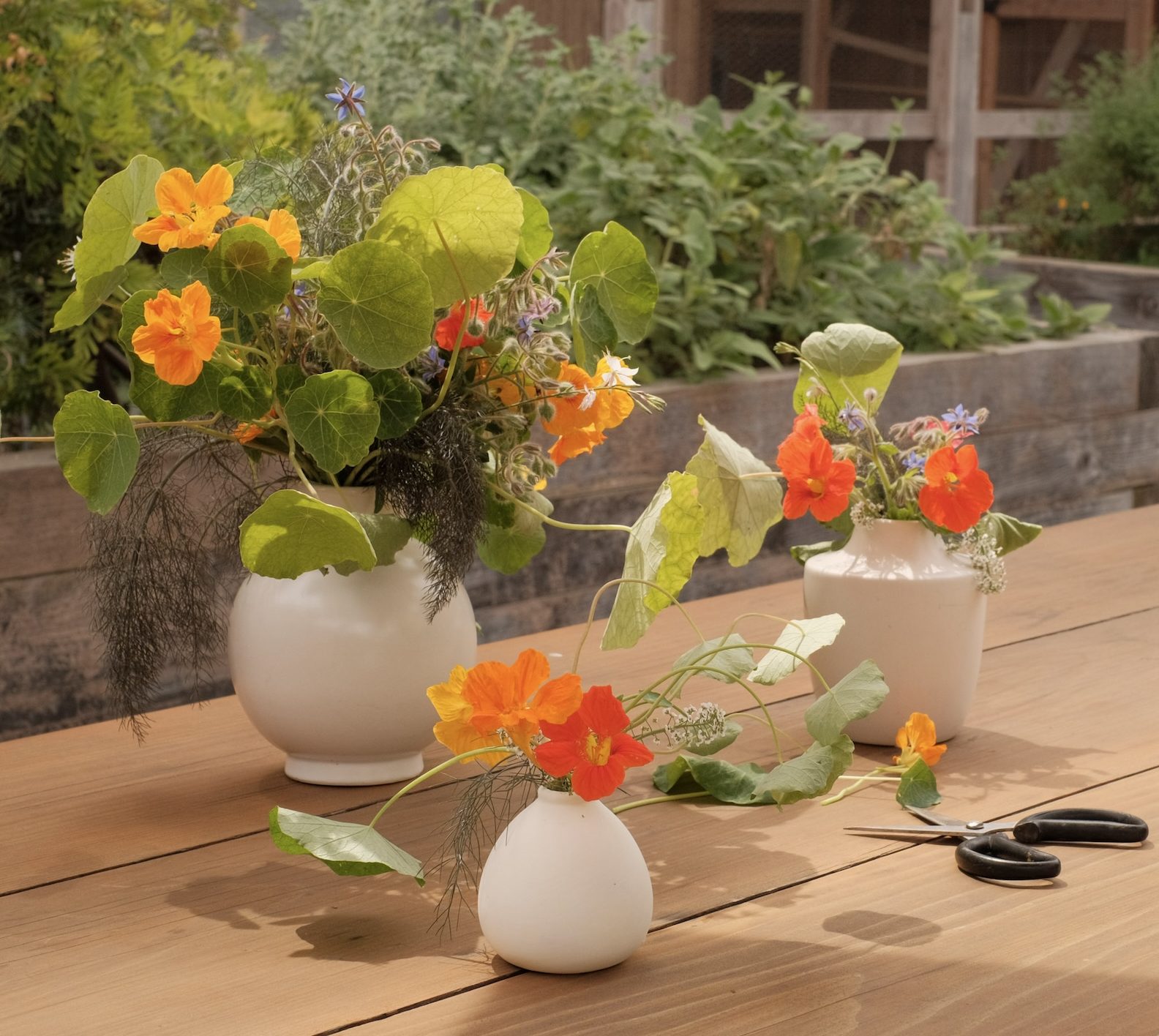 Three white vases in various shapes and sizes on an outdoor wooden table. Each vase has a unique arrangement of orange and red flowers.