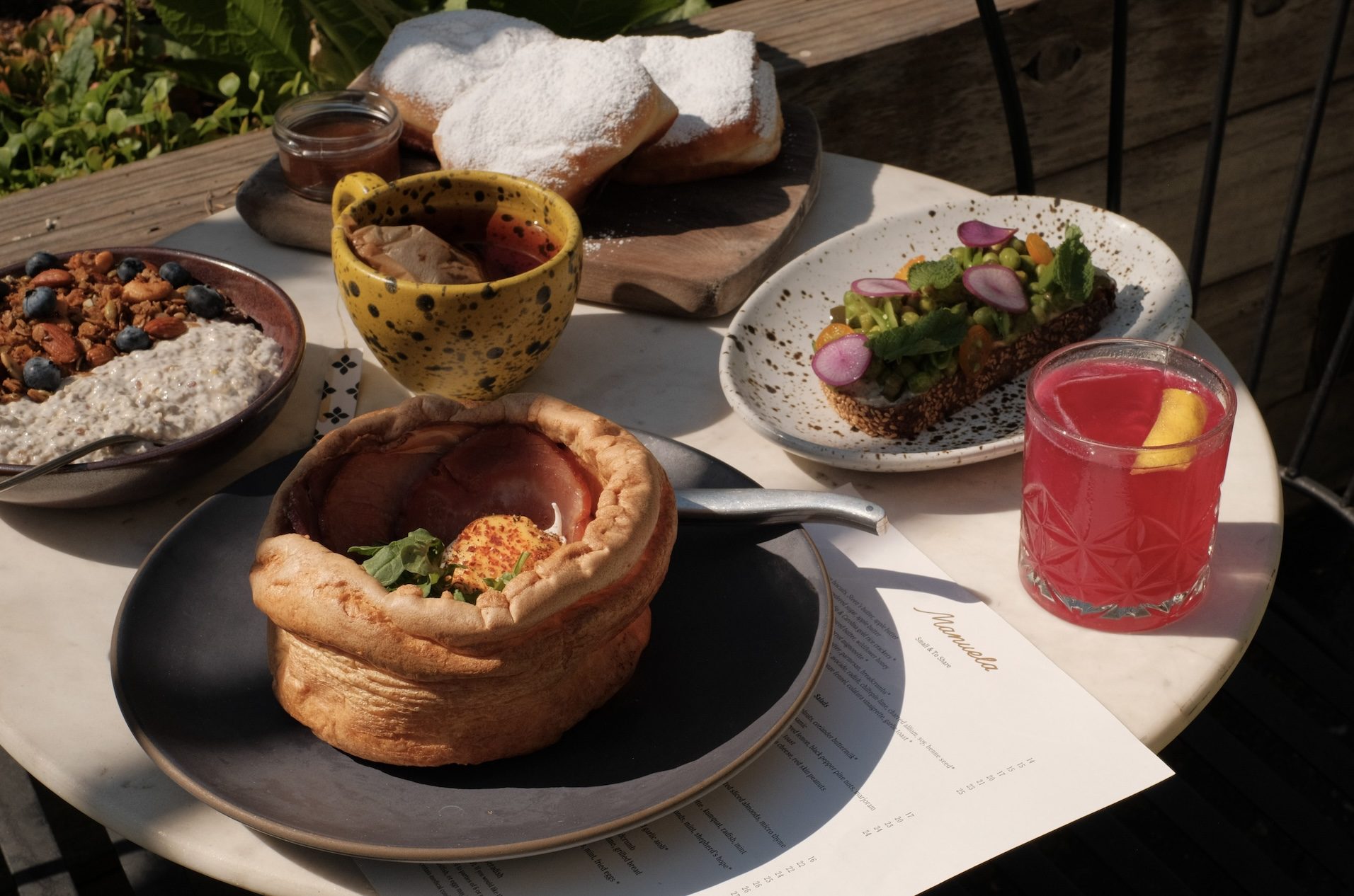 Brunch food on a white table including a large popover with eggs Benedict, an electric pink cocktail, and a granola bowl.