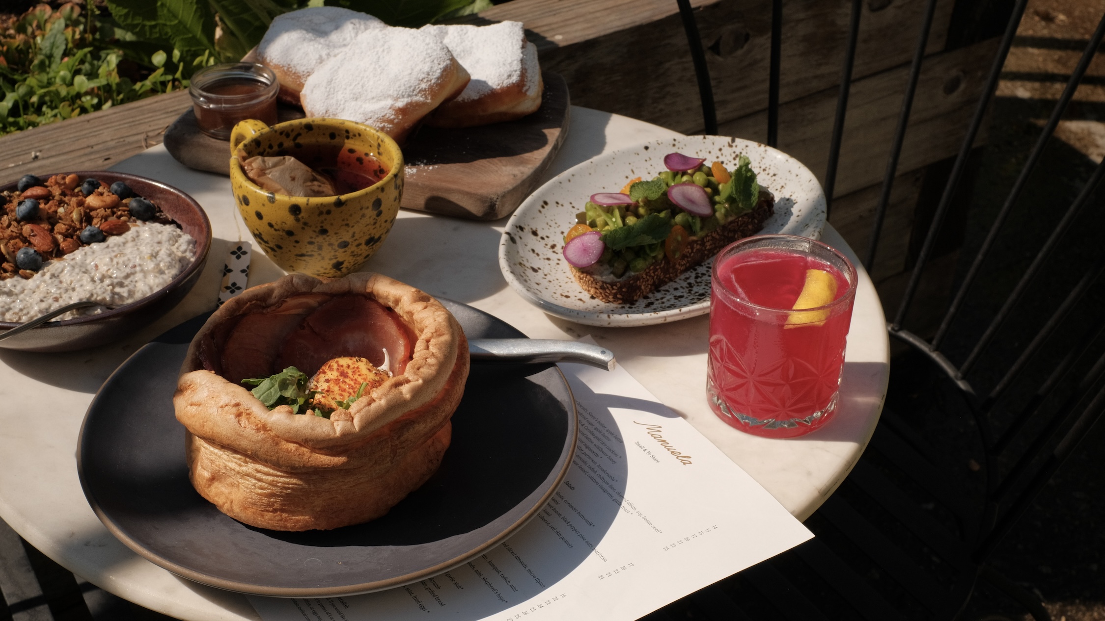 Brunch food on a white table including a large popover with eggs Benedict, an electric pink cocktail, and a granola bowl.