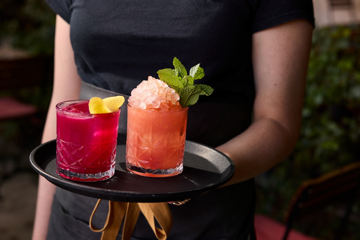 Server carrying a tray of two cocktails in a rocks glass. One is fuchsia with a yellow garnish, the other a peach tone with overflowing ice and a mint sprig.