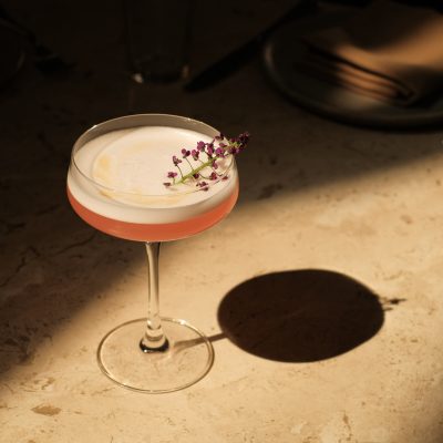 A pink cocktail in a coupe glass on a textured beige bar top with a flower stem garnish with small deep pink buds.