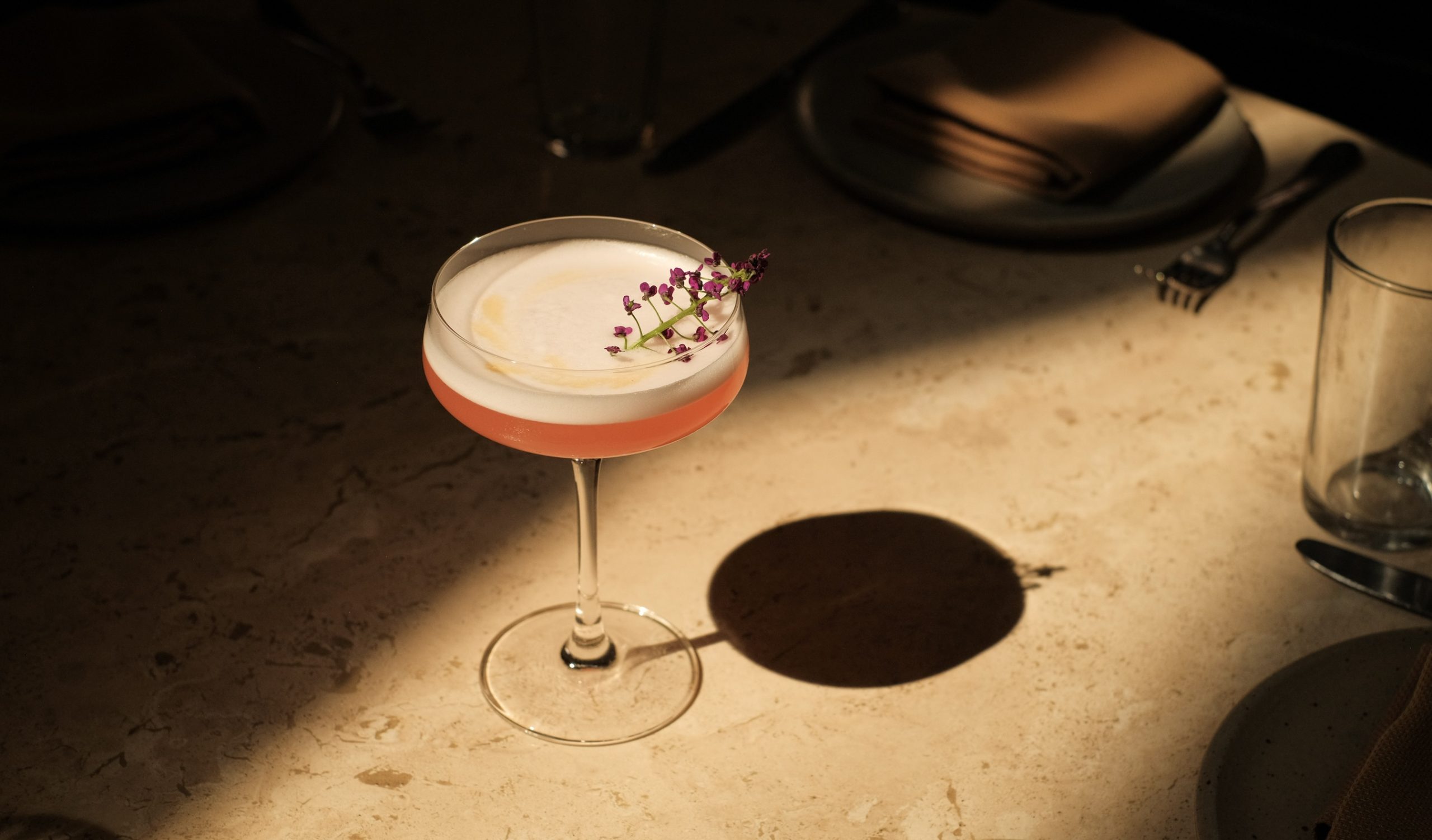 A pink cocktail in a coupe glass on a textured beige bar top with a flower stem garnish with small deep pink buds.