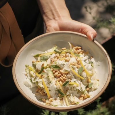 Hand holding a shallow white bowl with shaved asparagus, fennel, white cheese, and toped with pine nuts.