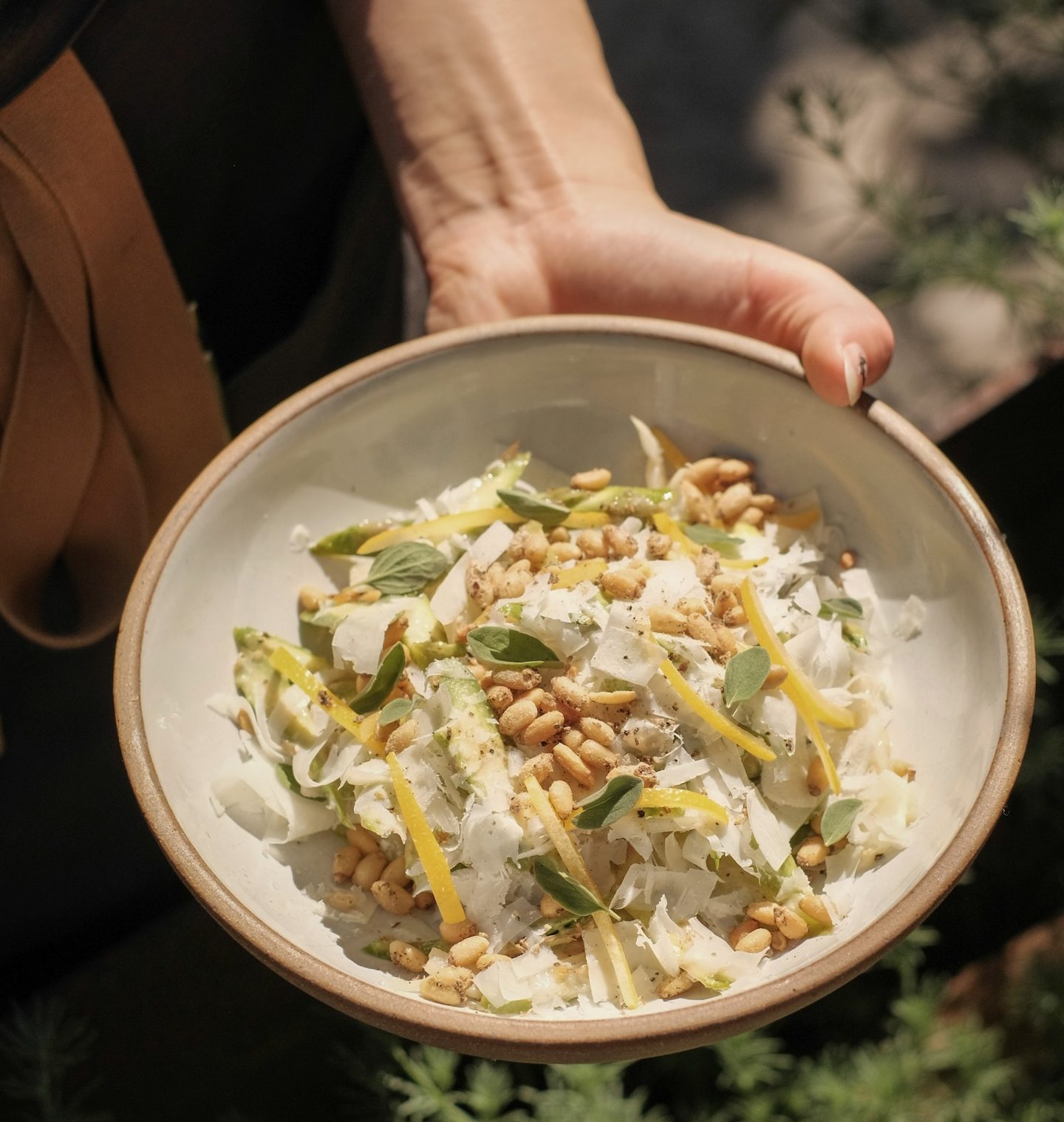 Hand holding a shallow white bowl with shaved asparagus, fennel, white cheese, and toped with pine nuts.