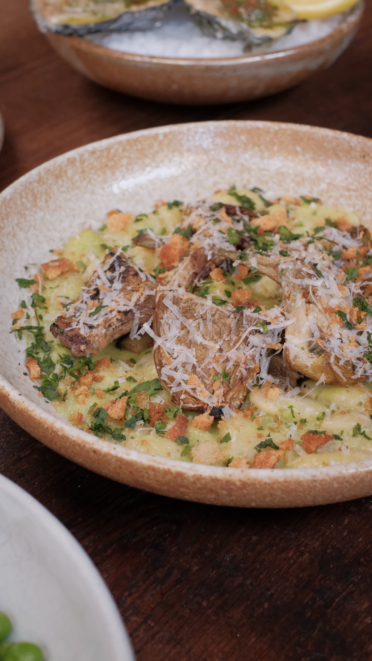 A close up of a round shallow bowl full of cooked white beams topped with roasted artichoke harts and garnished with a white cheese, green herbs, and bread crumbs. The dish is roasted artichokes, Lima beans, leek marscapone, Parmesan, duck fat breadcrumb