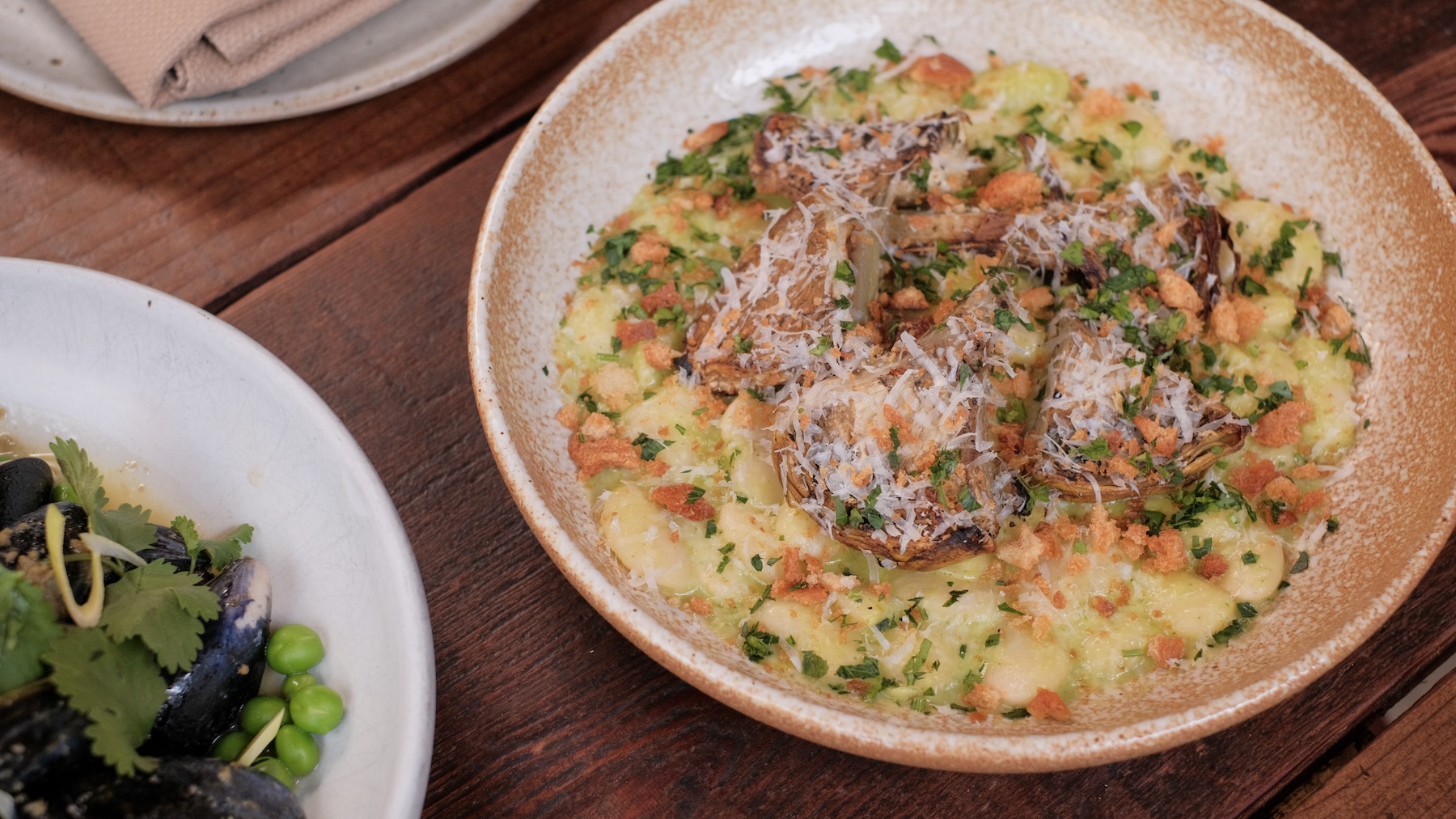 A round shallow bowl full of cooked white beams topped with roasted artichoke harts and garnished with a white cheese, green herbs, and bread crumbs. The dish is roasted artichokes, Lima beans, leek marscapone, Parmesan, duck fat breadcrumb
