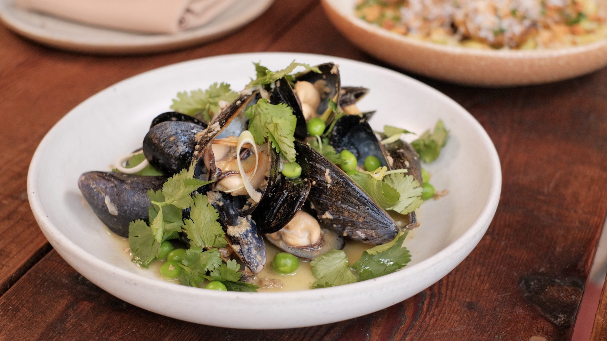A white, shallow bowl full of mussels and peas in a light broth, garnished with cilantro leaves.