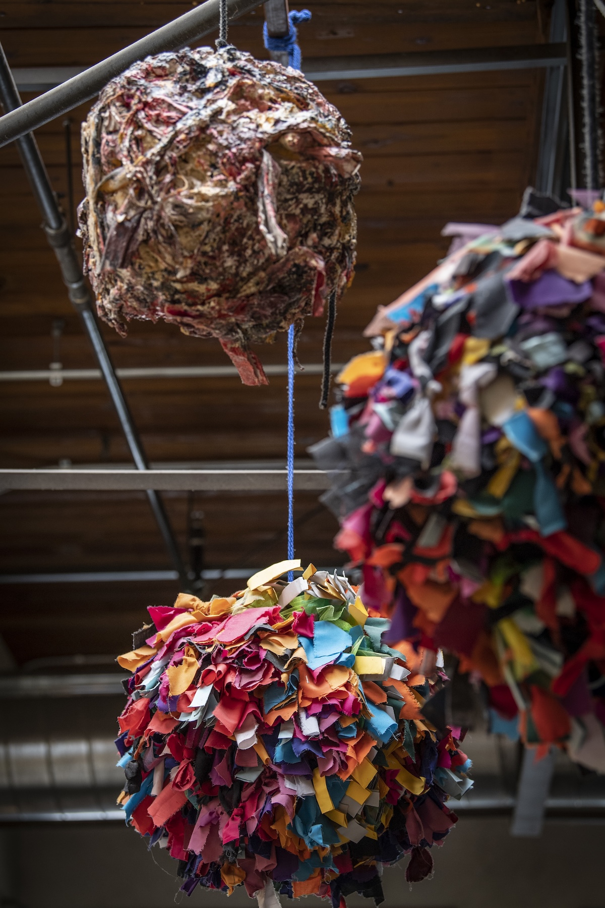 Three circular sculptures hanging from the ceiling . Two are made of countless pieces of color fabric and the other has a textured rock-like surface in more muted colors