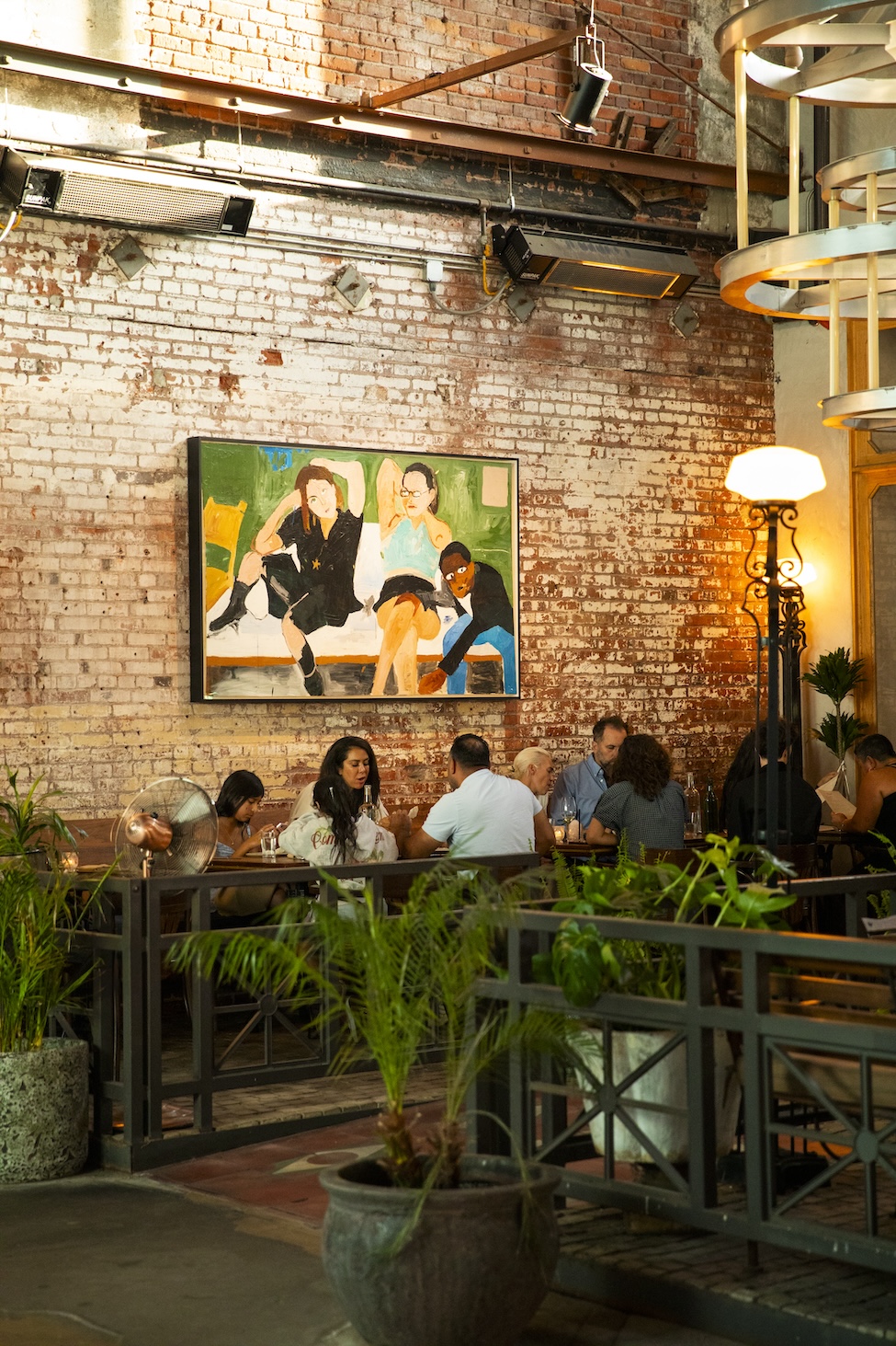 A colorful painting with blurred brush strokes of people talking together. The painting is hung on a brick wall with people sitting below it.