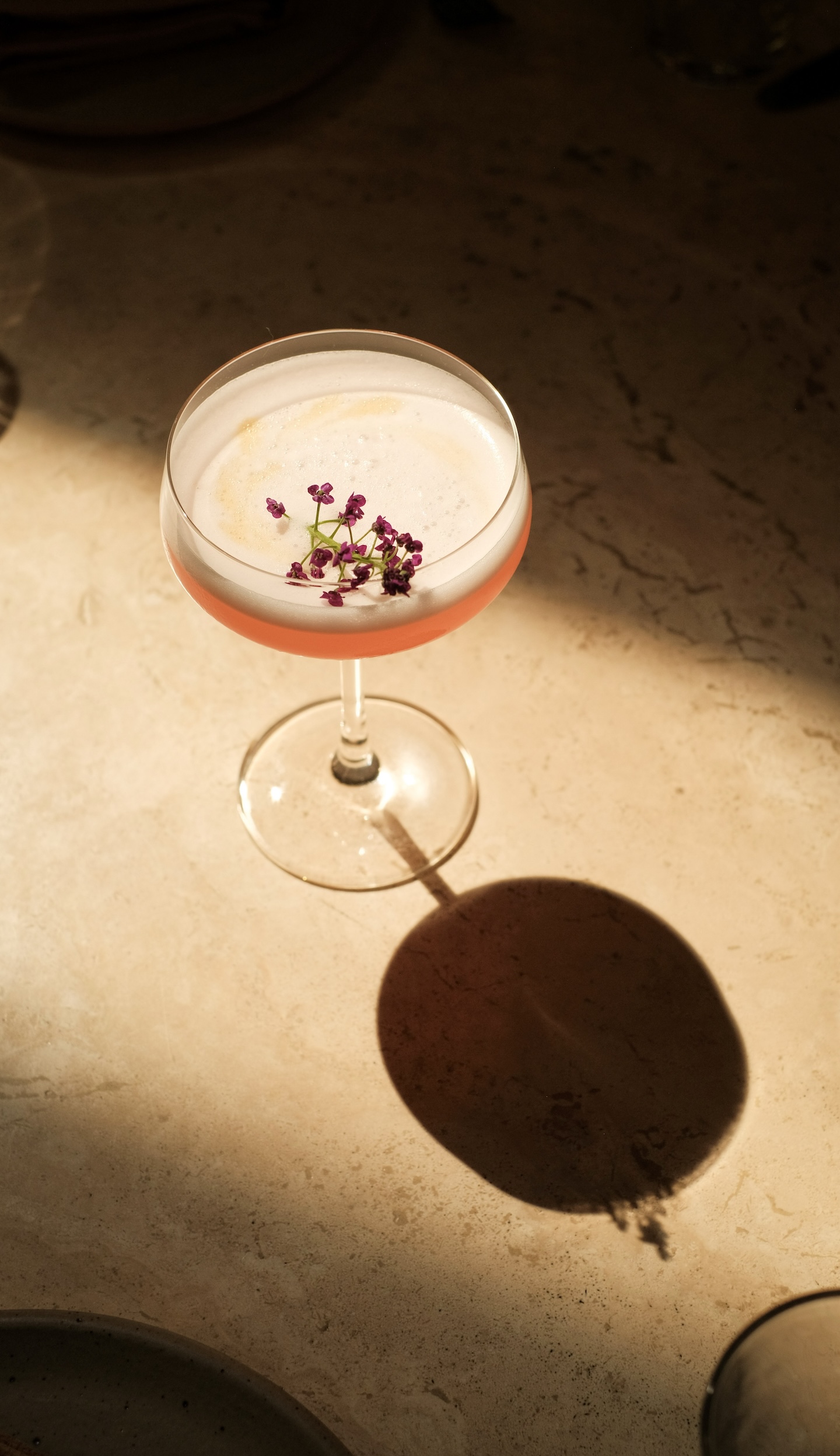 A pink cocktail in a coupe glass on a textured beige bar top with a flower stem garnish with small deep pink buds.