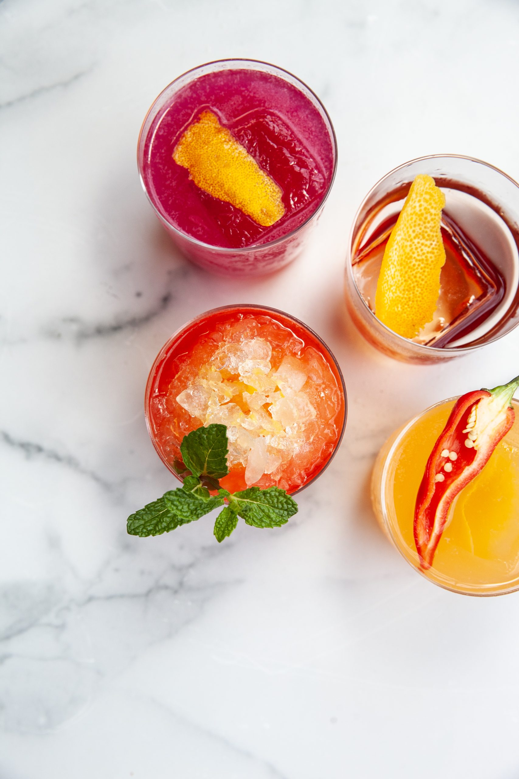 A grouping of brightly colored orange, red, and pink cocktails with various garnishes on a white marble top.