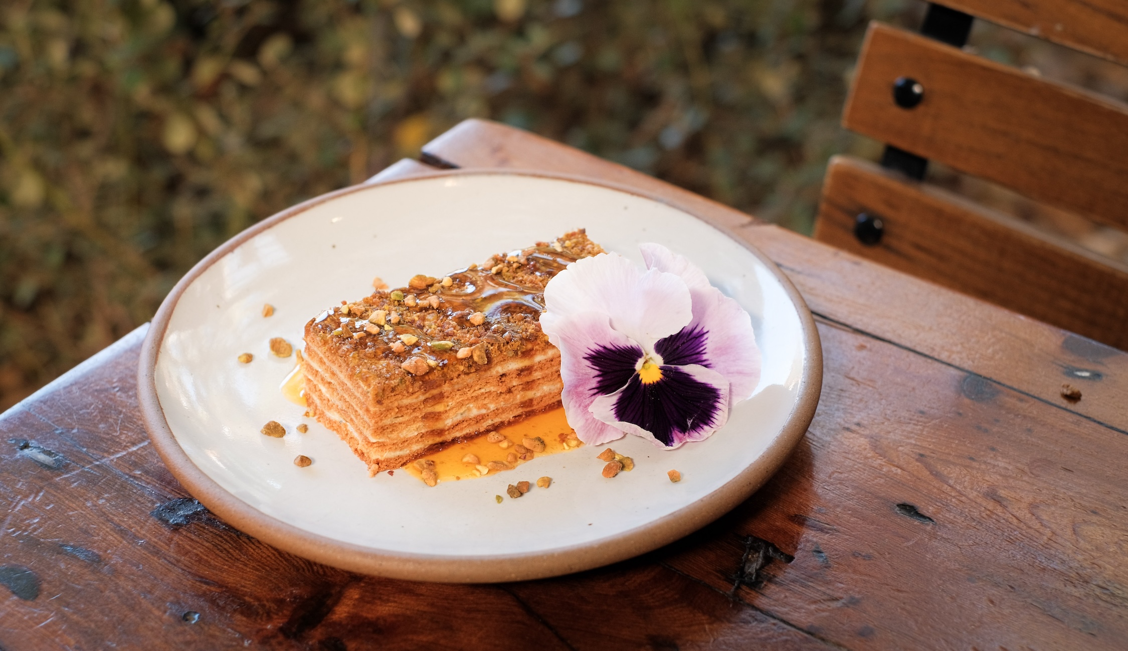 A layered cake with a syrup dripping off the top down the side. Nuts are sprinkled on top and it's garnished with a purple flower.