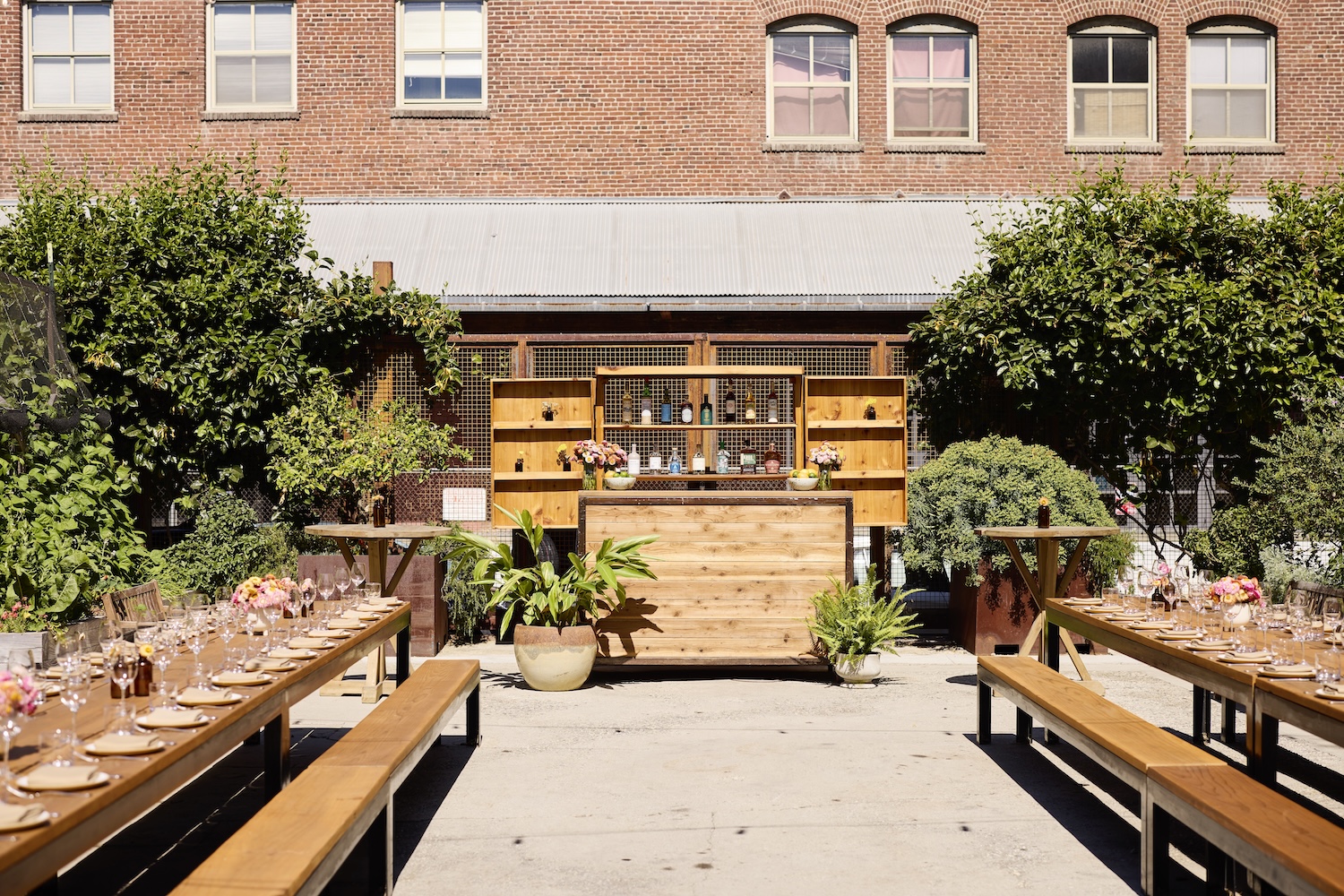 Two long tables set for dinner service in an outdoor garden area with a wooden bar framed in between them towards the back of the space.