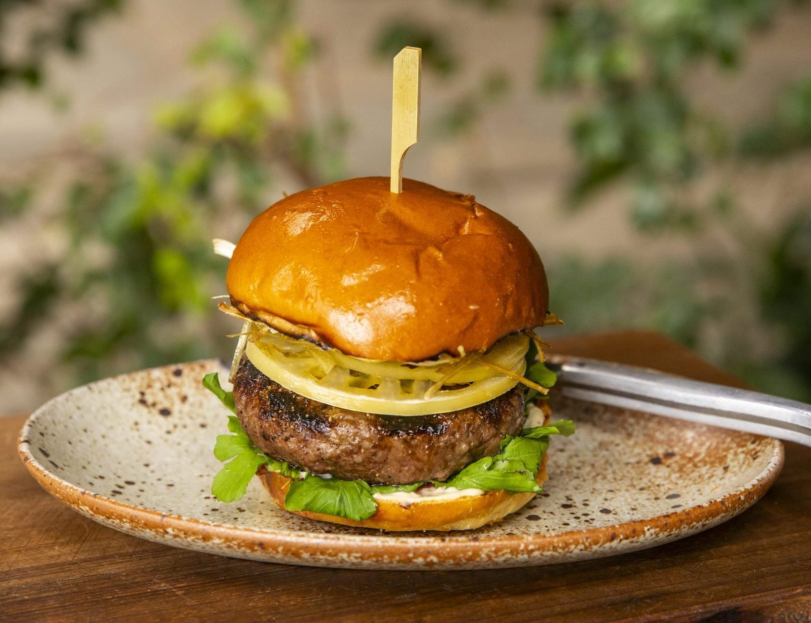Burger sitting on a textured plate with a knife behind it.The burger is dressed with green tomates and arugula, and this is a Venison burger on a brioche bun.