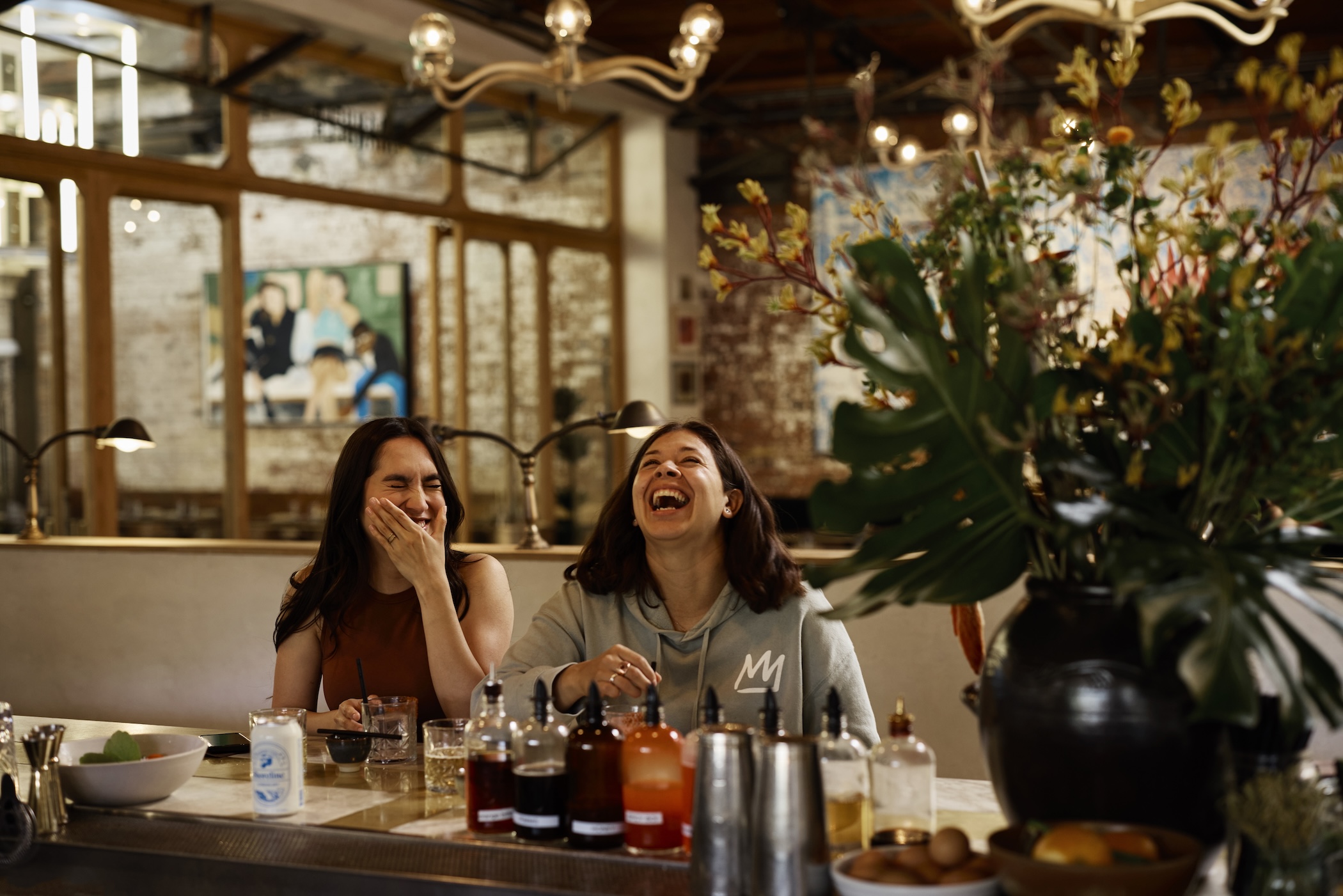 Two women laughing and enjoying drinks at a bar, with bottles and a vase of flowers in the foreground.