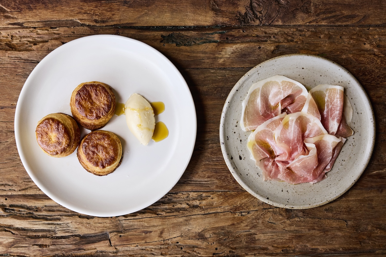Two plates on a wooden table: one with three golden biscuits and a dollop of butter, and the other with thin slices of cured ham.