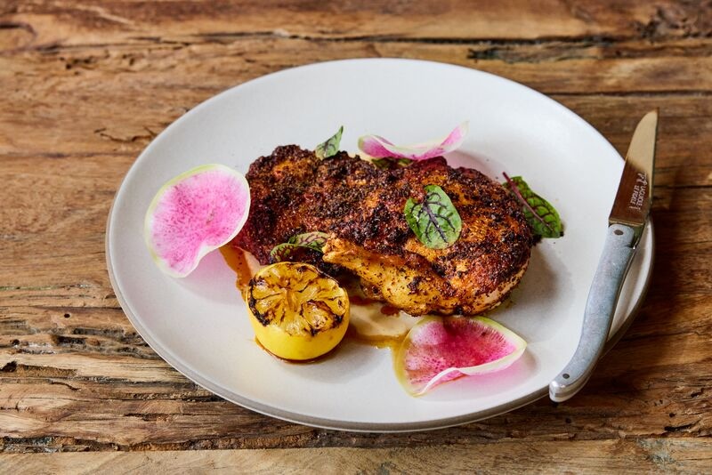 Seared chicken breast topped with herbs, served with grilled lemon and pink radish slices on a white plate.